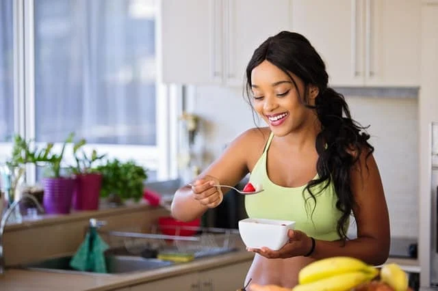 A woman in a green sports bra smiles while eating fruit from a white bowl in a bright kitchen with potted plants and bananas visible on the counter.