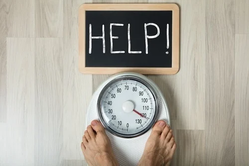 A person stands barefoot on a weighing scale, with a small chalkboard above displaying the word "HELP!" in large letters.