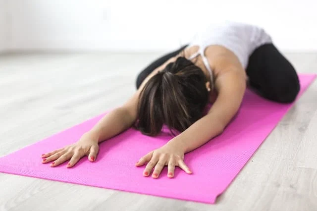 A person is practicing yoga on a pink mat indoors, stretching forward with arms extended and forehead touching the mat, wearing a white tank top and black pants.