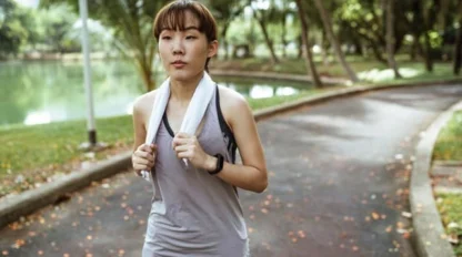A woman with a towel around her neck walks on a paved path in a park near a body of water, surrounded by trees.
