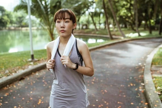A woman with a towel around her neck walks on a paved path in a park near a body of water, surrounded by trees.