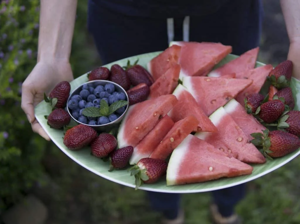 A person holds a large platter with watermelon slices, strawberries, and a small bowl of blueberries topped with a mint leaf.