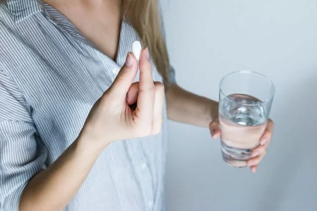 A person in a striped shirt holds a white pill in one hand and a glass of water in the other.