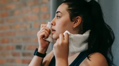 A woman with dark hair in a ponytail wipes her face with a white towel. She is wearing a black smartwatch and athletic clothing, standing in front of a brick wall.