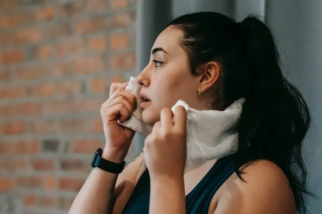 A woman with dark hair in a ponytail wipes her face with a white towel. She is wearing a black smartwatch and athletic clothing, standing in front of a brick wall.