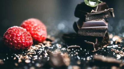 Two raspberries sit next to a stacked pile of dark chocolate pieces, garnished with a small green leaf, on a dark surface scattered with chocolate shavings.