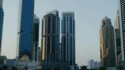 A waterfront cityscape with modern high-rise buildings and skyscrapers at sunset, reflected in the calm water. A curved metal railing lines the walkway in the foreground.