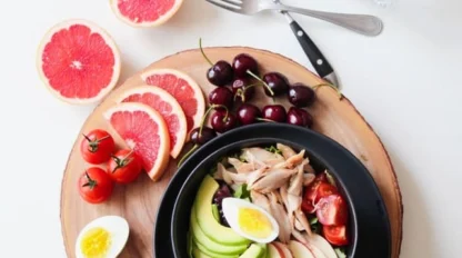A wooden platter with sliced grapefruit, cherries, tomatoes, a hard-boiled egg, and a black bowl filled with avocado, shredded chicken, tomato, and egg slices. Spoons, forks, and a glass are in the background.
