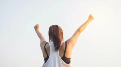 A person with long hair in a ponytail, wearing a sleeveless top, stands outdoors with arms raised triumphantly against a bright, clear sky.