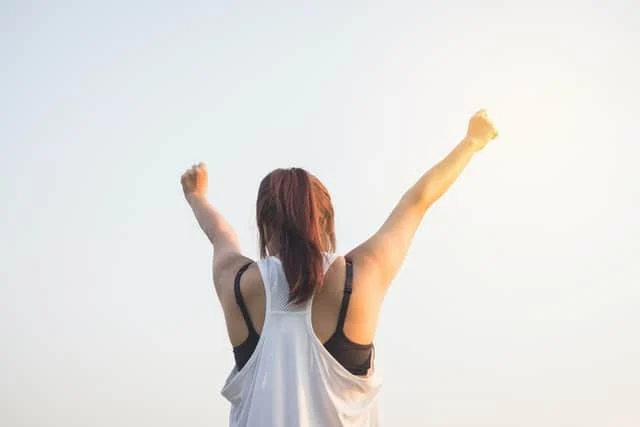 A person with long hair in a ponytail, wearing a sleeveless top, stands outdoors with arms raised triumphantly against a bright, clear sky.