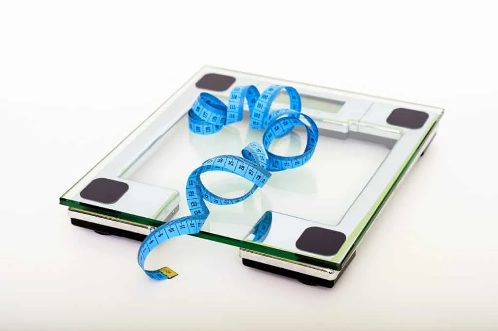 A glass digital bathroom scale with a coiled blue measuring tape placed on top, set against a plain white background.
