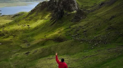 A person in a red jacket stands on a grassy hill, facing and waving toward a dramatic rock formation in a green, rolling landscape with distant mountains and water under a cloudy sky.