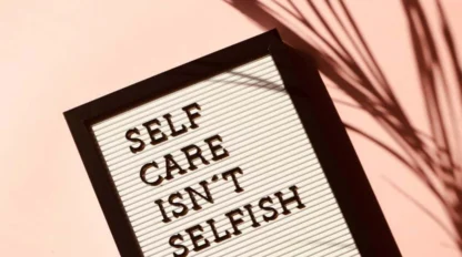 A letter board with black text reads "SELF CARE ISN'T SELFISH" on a pink background with a shadow of a plant.