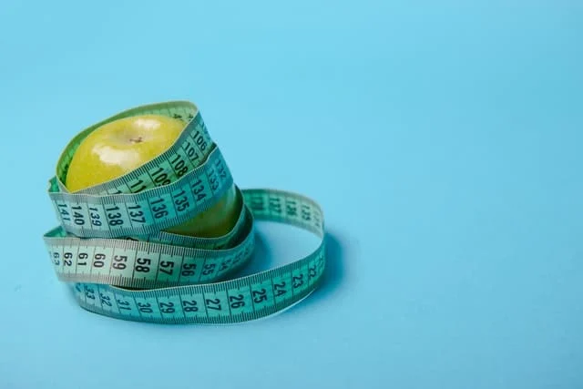 A green apple with a measuring tape wrapped around it, set against a plain blue background.