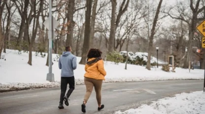 Two people jogging along a road in a snowy park, surrounded by bare trees and snow-covered ground.