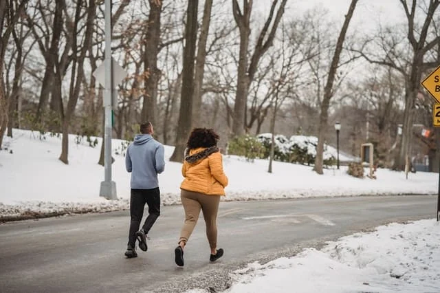 Two people jogging along a road in a snowy park, surrounded by bare trees and snow-covered ground.