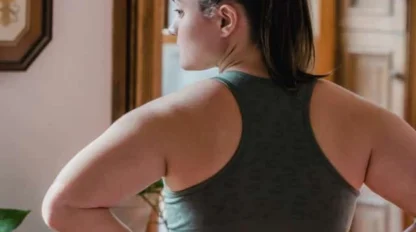 A woman in athletic wear stands indoors with her hands on her lower back, facing away from the camera.