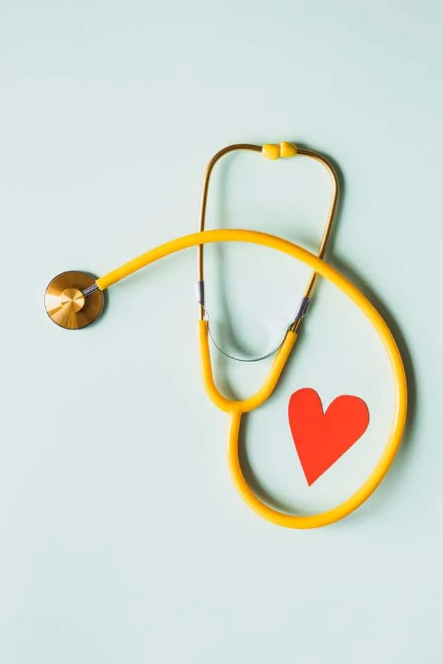 A yellow stethoscope is arranged on a light surface next to a red paper heart shape.