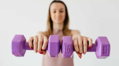 A person with red nail polish holds two purple dumbbells toward the camera, slightly out of focus, against a plain light background.