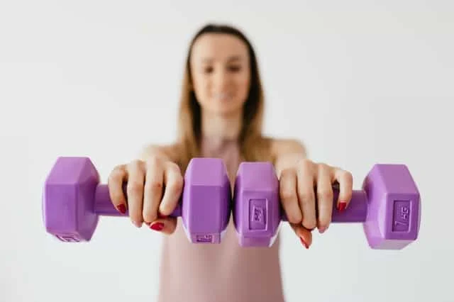 A person with red nail polish holds two purple dumbbells toward the camera, slightly out of focus, against a plain light background.