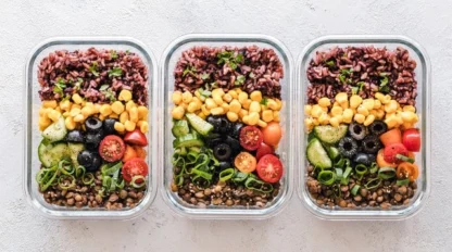 Three glass containers filled with neatly arranged brown rice, corn, black olives, cucumbers, cherry tomatoes, lentils, and green onions on a light surface.