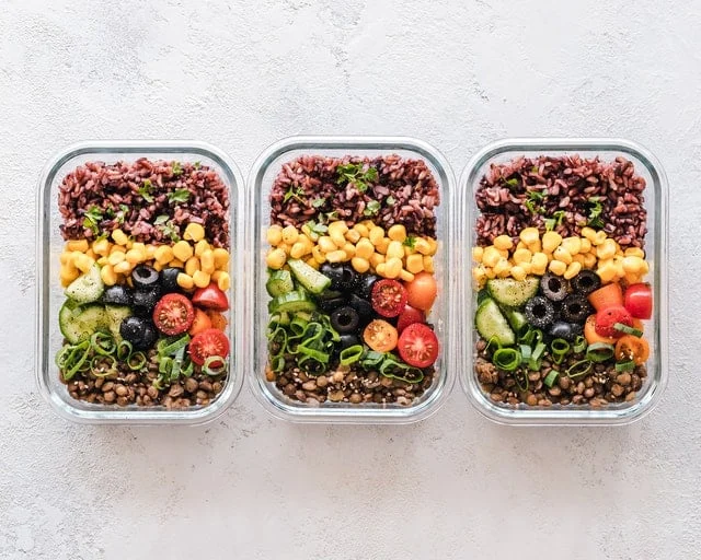 Three glass containers filled with neatly arranged brown rice, corn, black olives, cucumbers, cherry tomatoes, lentils, and green onions on a light surface.