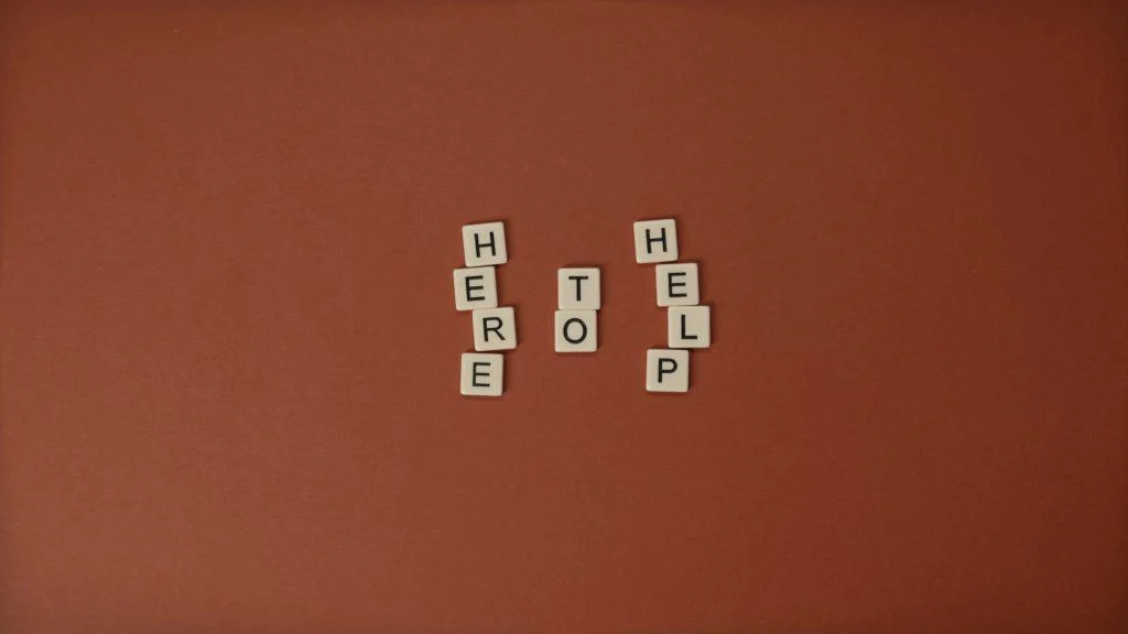 Scrabble tiles arranged on a brown background spell out the phrase "HERE TO HELP" in three vertical columns.
