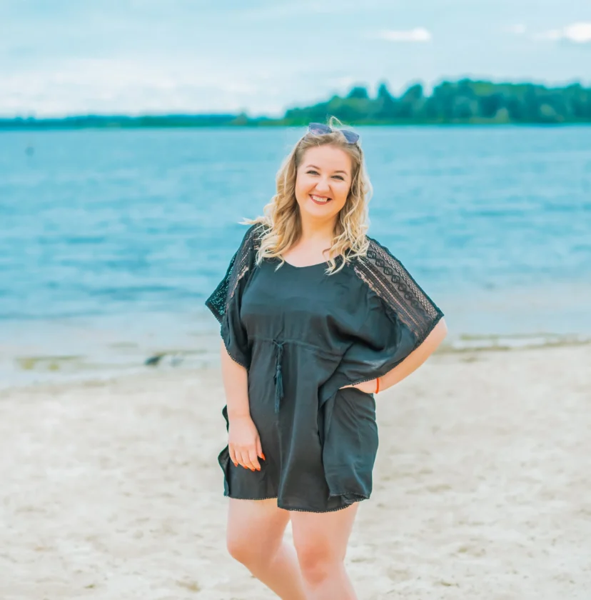 Person in a black outfit stands barefoot on sandy beach, smiling. Calm water and trees visible in the background under a cloudy sky.