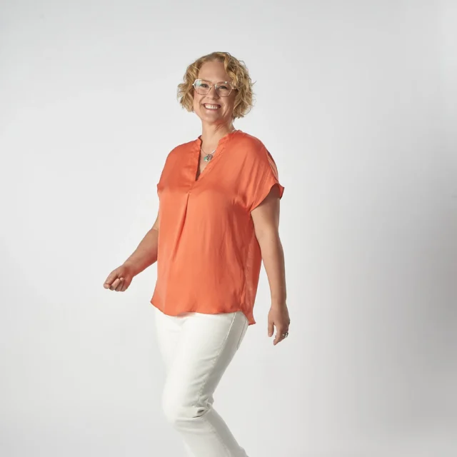 A person wearing an orange blouse and white pants stands against a plain background, smiling.