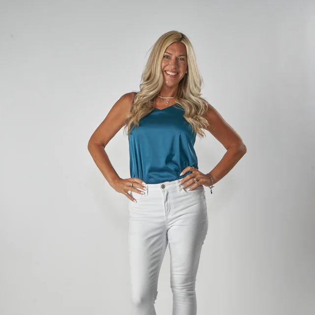 A woman with long blonde hair wearing a blue sleeveless top and white pants smiles at the camera against a plain background.