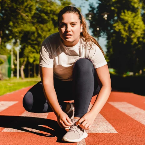 A person tying their shoe on a red running track in a park during the day, surrounded by green trees and grass.
