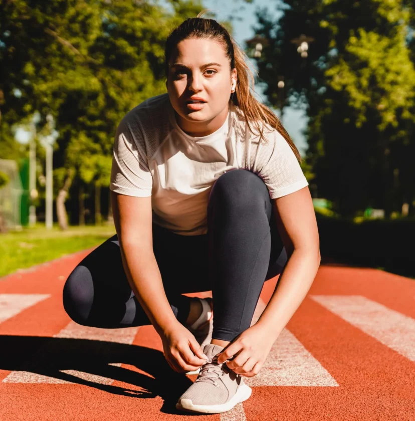 A person tying their shoe on a red running track in a park during the day, surrounded by green trees and grass.