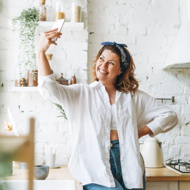 A person in a white shirt and blue headband takes a selfie in a bright kitchen with white brick walls and various kitchen items.