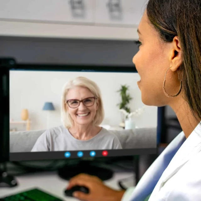 A woman in a white coat has a video call with another woman displayed on her computer monitor, both appearing engaged in conversation.