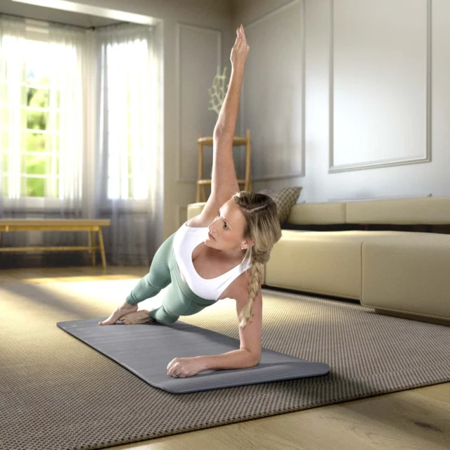 A woman performs a side plank on a yoga mat in a bright living room with modern decor and large windows.