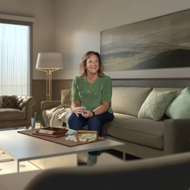 A woman sits on a sofa in a living room, smiling, with a board game and remote control on the coffee table in front of her.