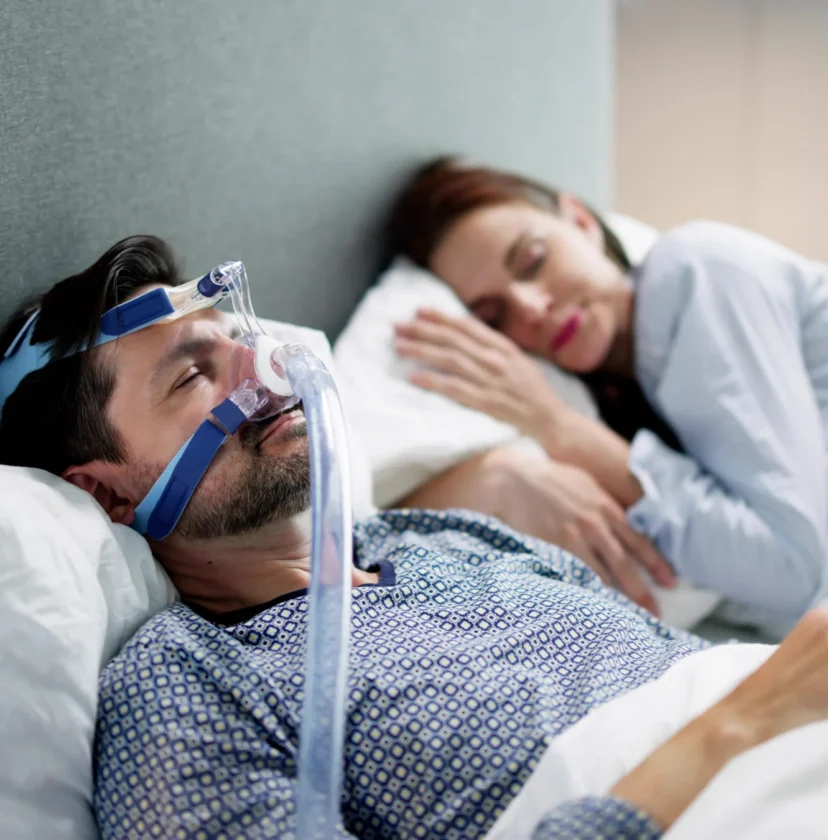 A man wearing a CPAP mask sleeps in bed next to a woman who is also sleeping.