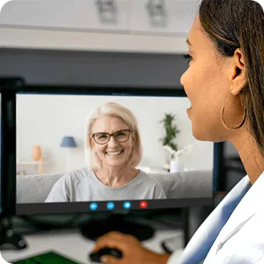 A woman participates in a video call with another woman shown smiling on a computer screen in a home or office setting.