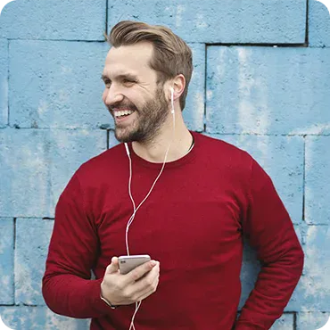 Man in a red sweater standing against a blue brick wall, smiling while holding a smartphone and wearing white earphones.