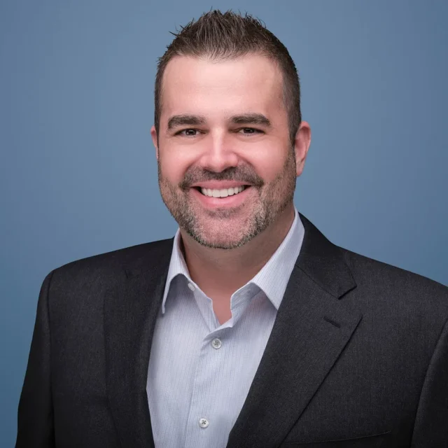 A man with short dark hair and a trimmed beard, wearing a dark suit jacket and light dress shirt, smiles in front of a plain blue background.