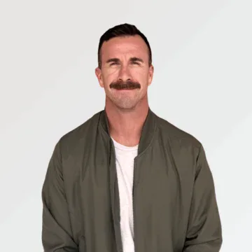 Man with short brown hair and a mustache wearing a white t-shirt and an olive green jacket standing against a plain light background.