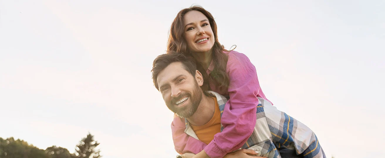 A man gives a woman a piggyback ride outdoors. Both are smiling and looking at the camera, with trees and sky in the background.