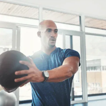 A man in athletic wear holds a medicine ball with both hands in a gym, appearing focused during a workout. Large windows and gym equipment are visible in the background.