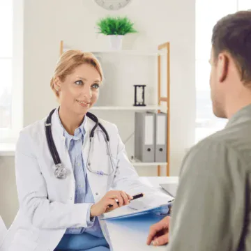 A female doctor in a white coat talks to a male patient across a desk in a bright medical office.