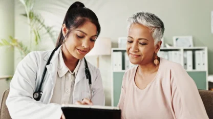 A female doctor in a white coat shows information on a tablet to an older woman with short gray hair. Both are smiling and sitting in a bright, modern office with shelves and plants in the background.