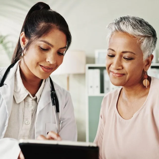 A female doctor in a white coat shows information on a tablet to an older woman with short gray hair. Both are smiling and sitting in a bright, modern office with shelves and plants in the background.