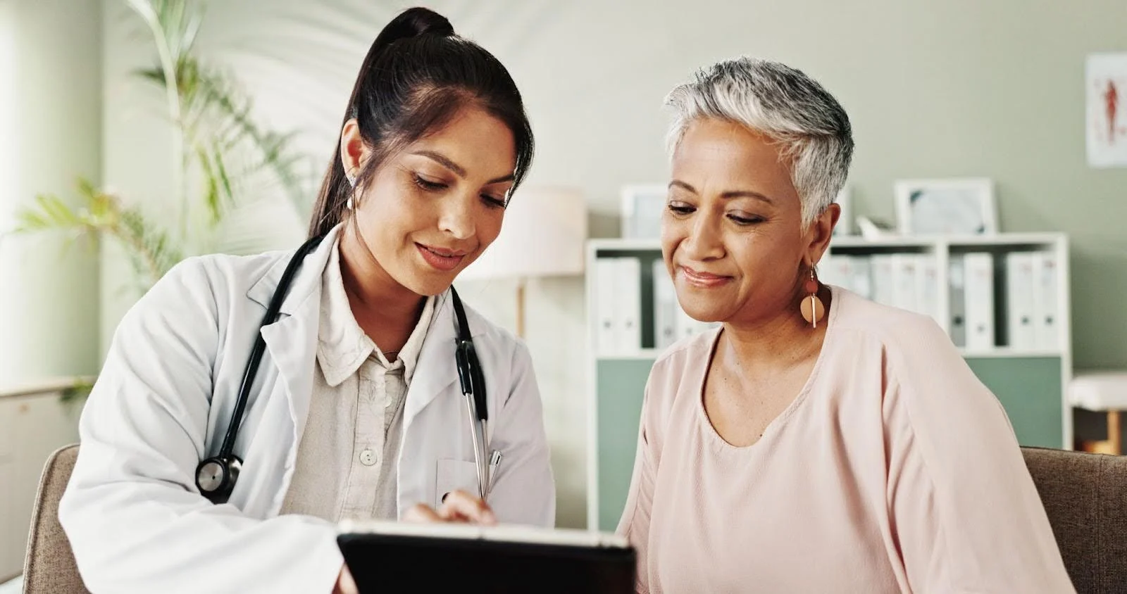 A female doctor in a white coat shows information on a tablet to an older woman with short gray hair. Both are smiling and sitting in a bright, modern office with shelves and plants in the background.