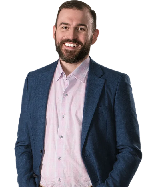 A man with a beard smiles, wearing a light pink button-up shirt and a blue blazer, standing against a white background.