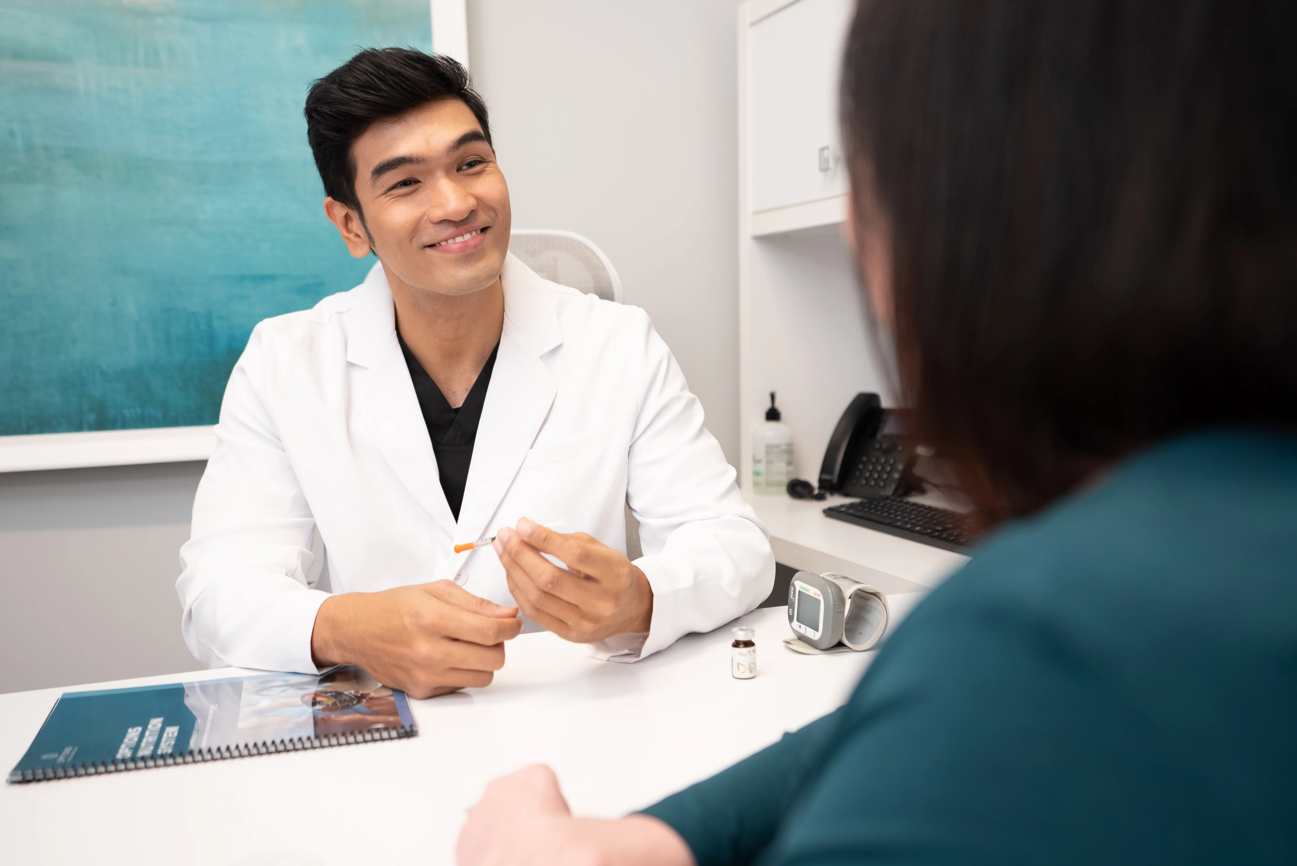 A female doctor wearing a white coat gently touches the shoulder of a smiling woman in an orange blouse as they talk in a medical office.