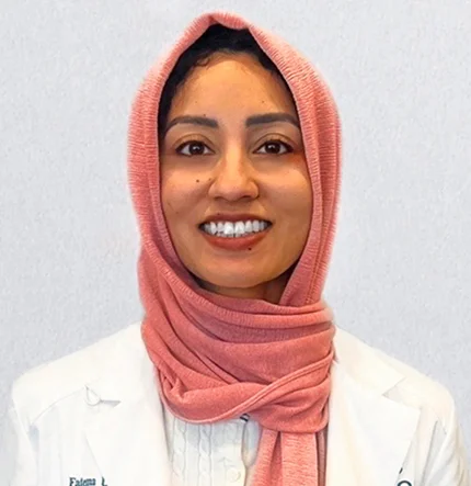 A woman wearing a light pink hijab and a white lab coat smiles at the camera, standing against a plain light background.